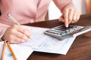 A woman sits at a table with a calculator, documents, and a pen, focused on her work.